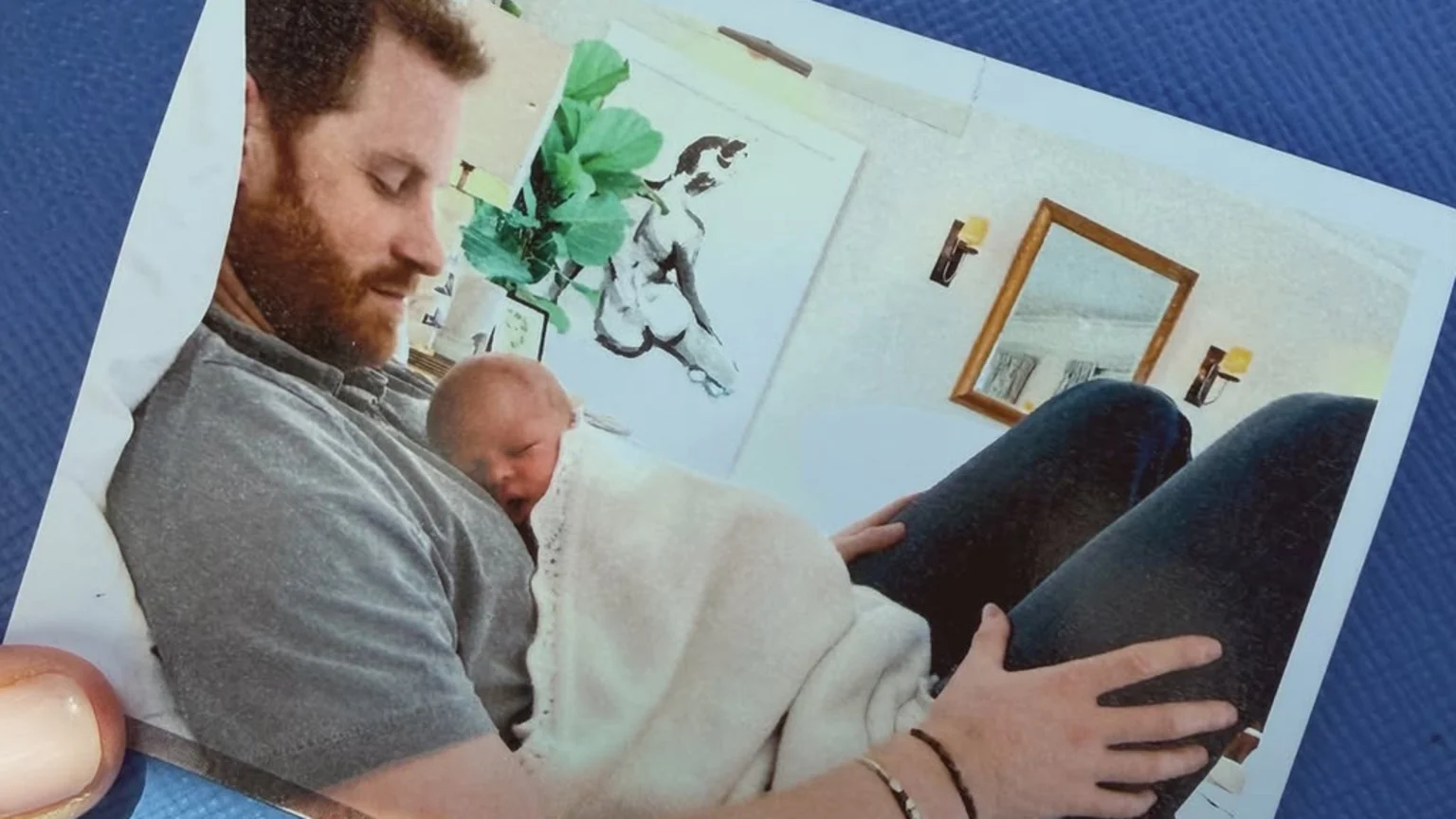 A bearded man in a grey shirt gently holds a sleeping baby wrapped in a white blanket, while a hand with bracelets rests on his leg; artwork and a mirror adorn the background.