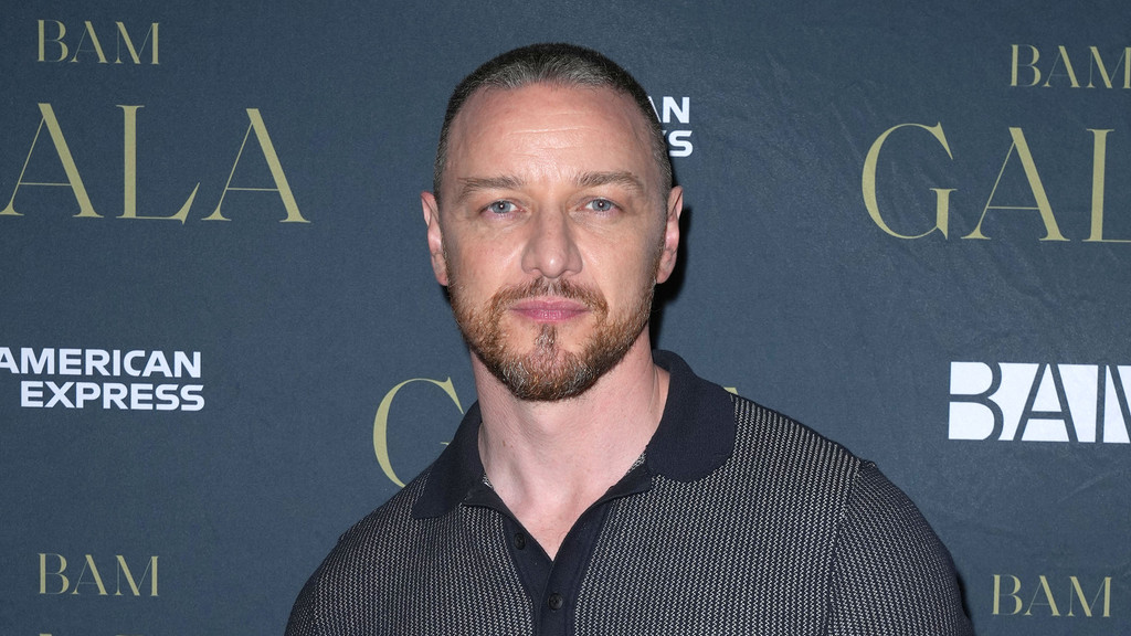 James McAvoy, with short hair and a beard, wears a dark patterned polo shirt and looks forward against a dark backdrop with "BAM GALA" and "AMERICAN EXPRESS" branding.