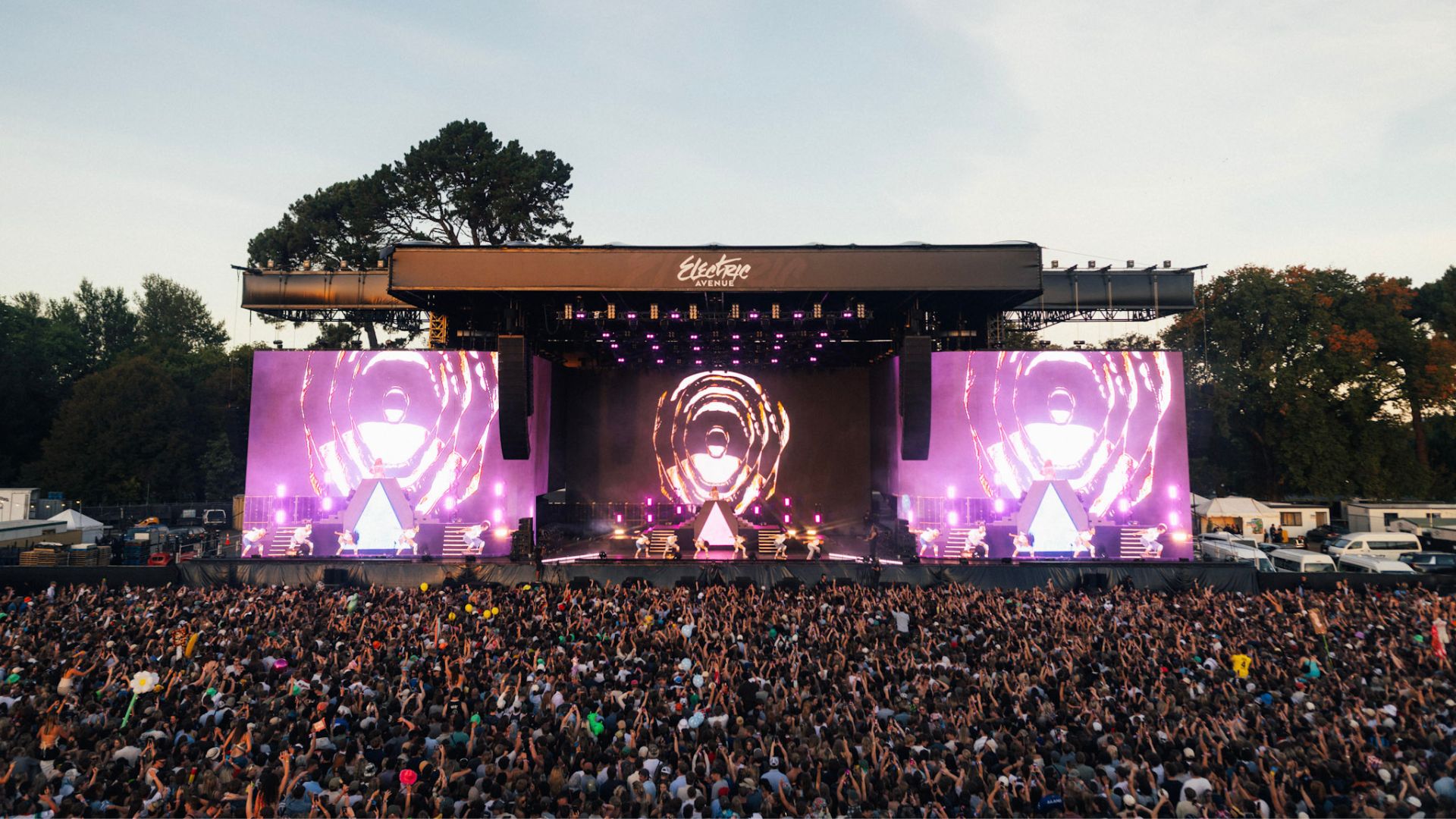 A wide shot of an outdoor concert at Electric Avenue, showing a stage with large purple screens displaying visuals, performers, and a massive audience with many hands raised.