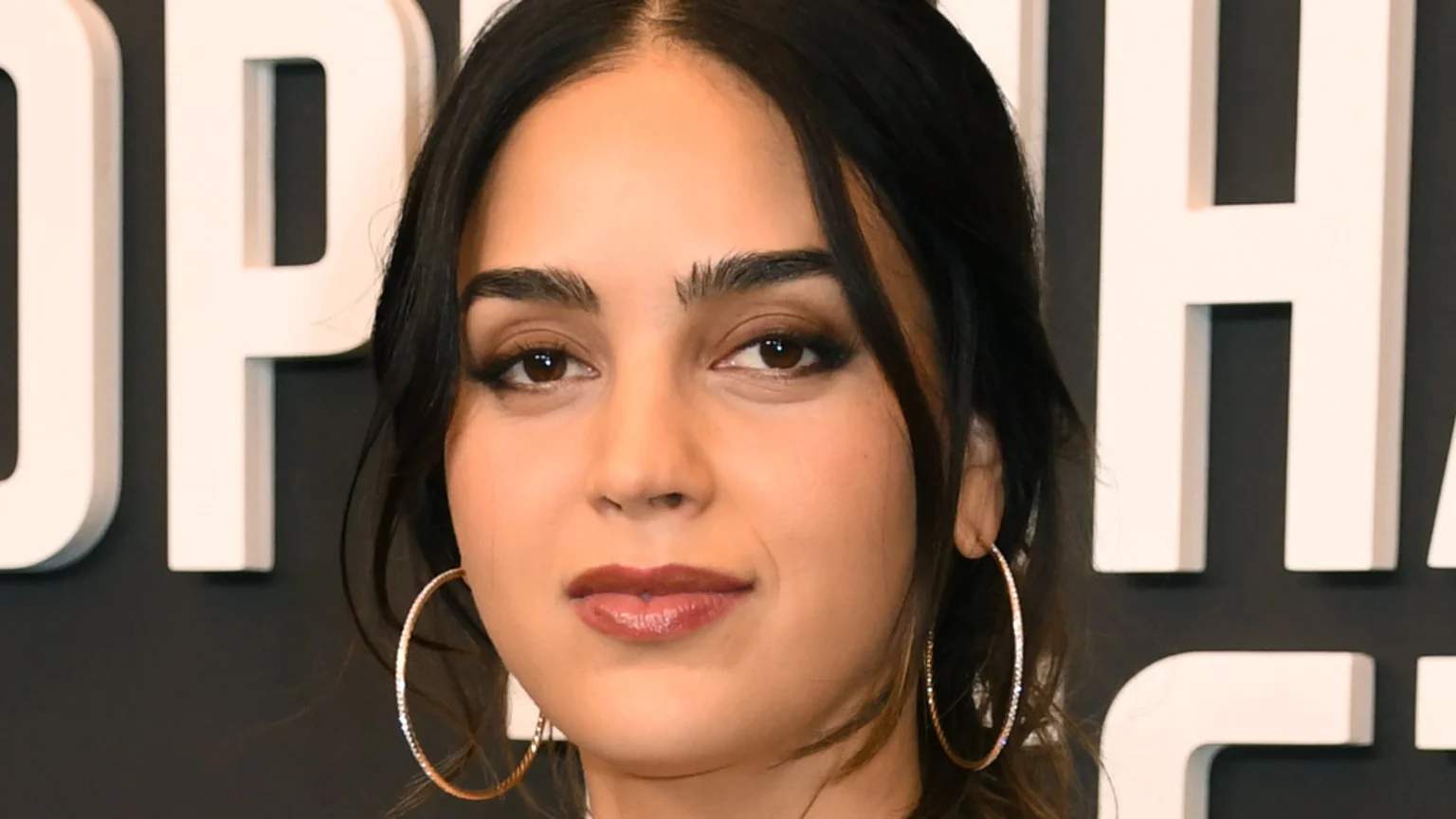 Close-up portrait of a woman with dark hair, brown eyes, and full eyebrows, wearing large textured gold hoop earrings and glossy reddish-brown lipstick, against a background of white dimensional letters.