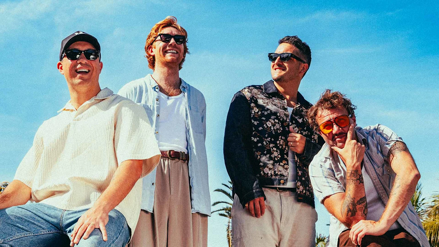 Four men, likely a band, pose outdoors under a bright blue sky, all wearing sunglasses and smiling or smirking in casual attire.
