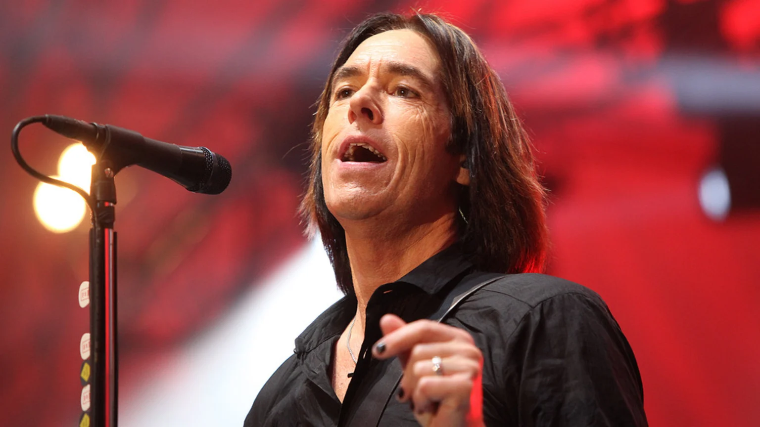 A male singer with long brown hair and a black shirt sings into a microphone on a red-lit stage, showing a ring and black nail polish.
