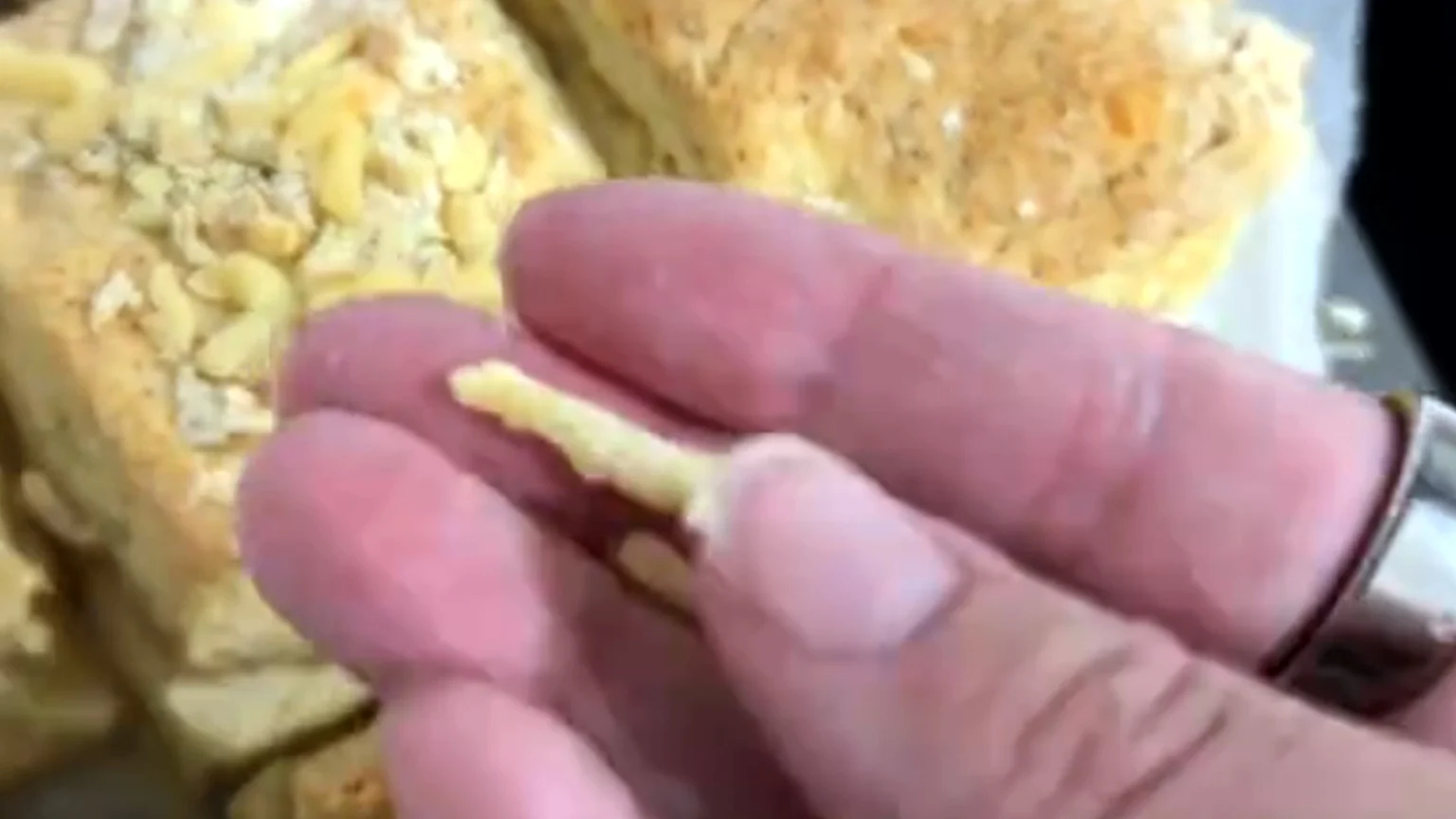 A hand with a silver ring holds a small, firm, yellowish-white strip of food, with out-of-focus baked goods topped with similar pieces visible in the background.