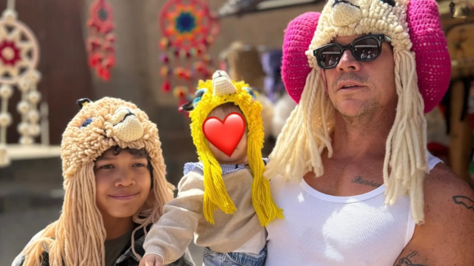 Diplo in sunglasses and a ram hat holds a baby, whose face is covered by a heart emoji, wearing a unicorn hat, next to a boy in a lion hat.