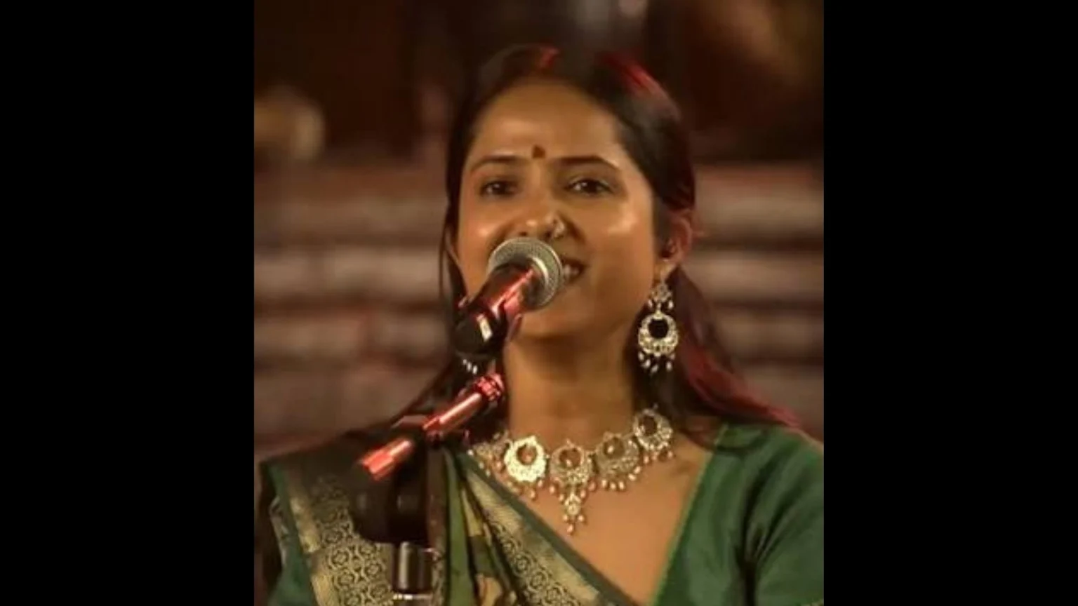 A woman in a green sari and traditional Indian jewelry, including a nose ring and bindi, smiles while singing into a microphone on a dimly lit stage.
