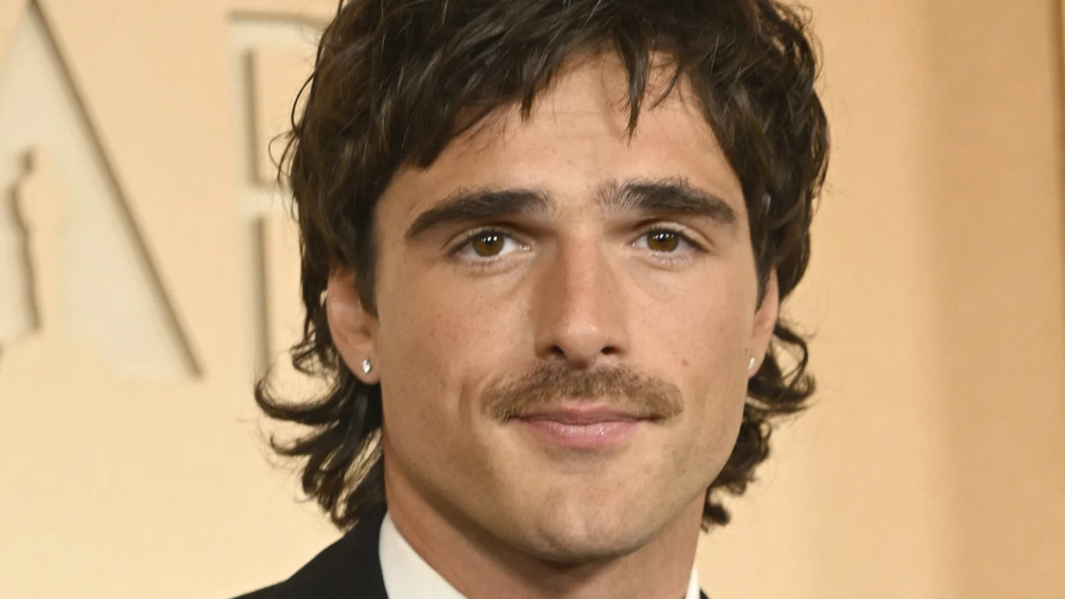 Close-up of a man with dark, wavy hair, a prominent mustache, brown eyes, and stud earrings, wearing a suit and smiling faintly, against a blurred, light background.