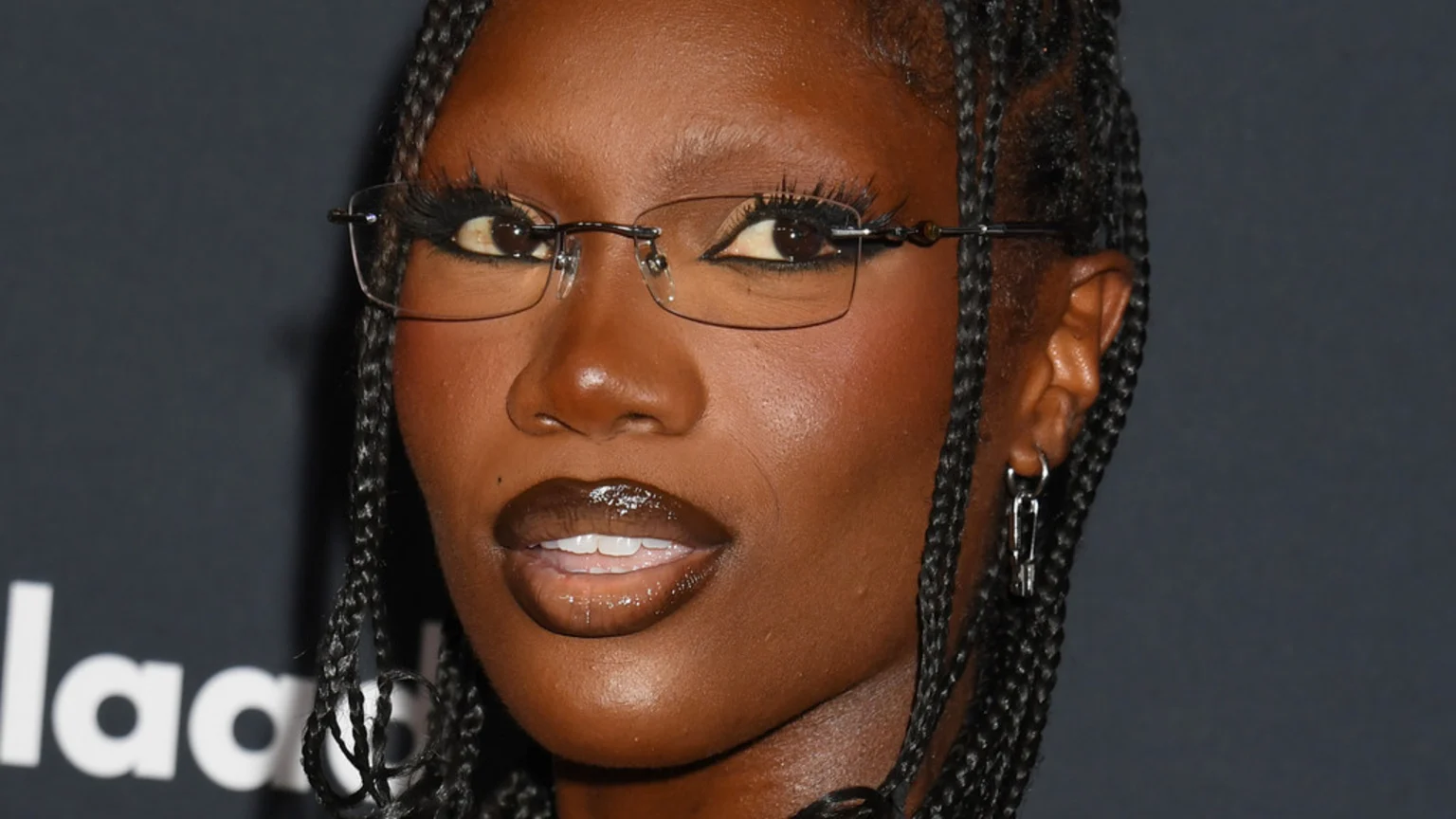 A close-up of a dark-skinned woman with braided hair, wearing frameless rectangular glasses, long eyelashes, and glossy dark reddish-brown lipstick, with a partial "glaa" logo in the background.