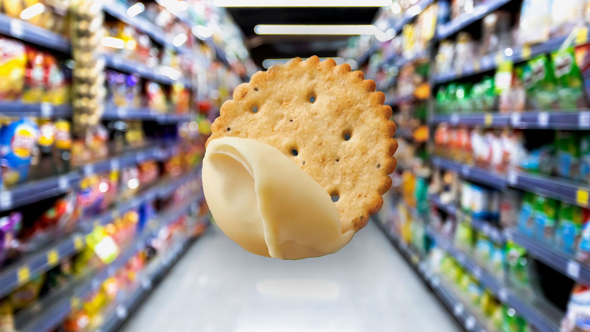 A round, scalloped cracker with a light-colored creamy spread floats in front of a blurred grocery store aisle filled with packaged snacks.