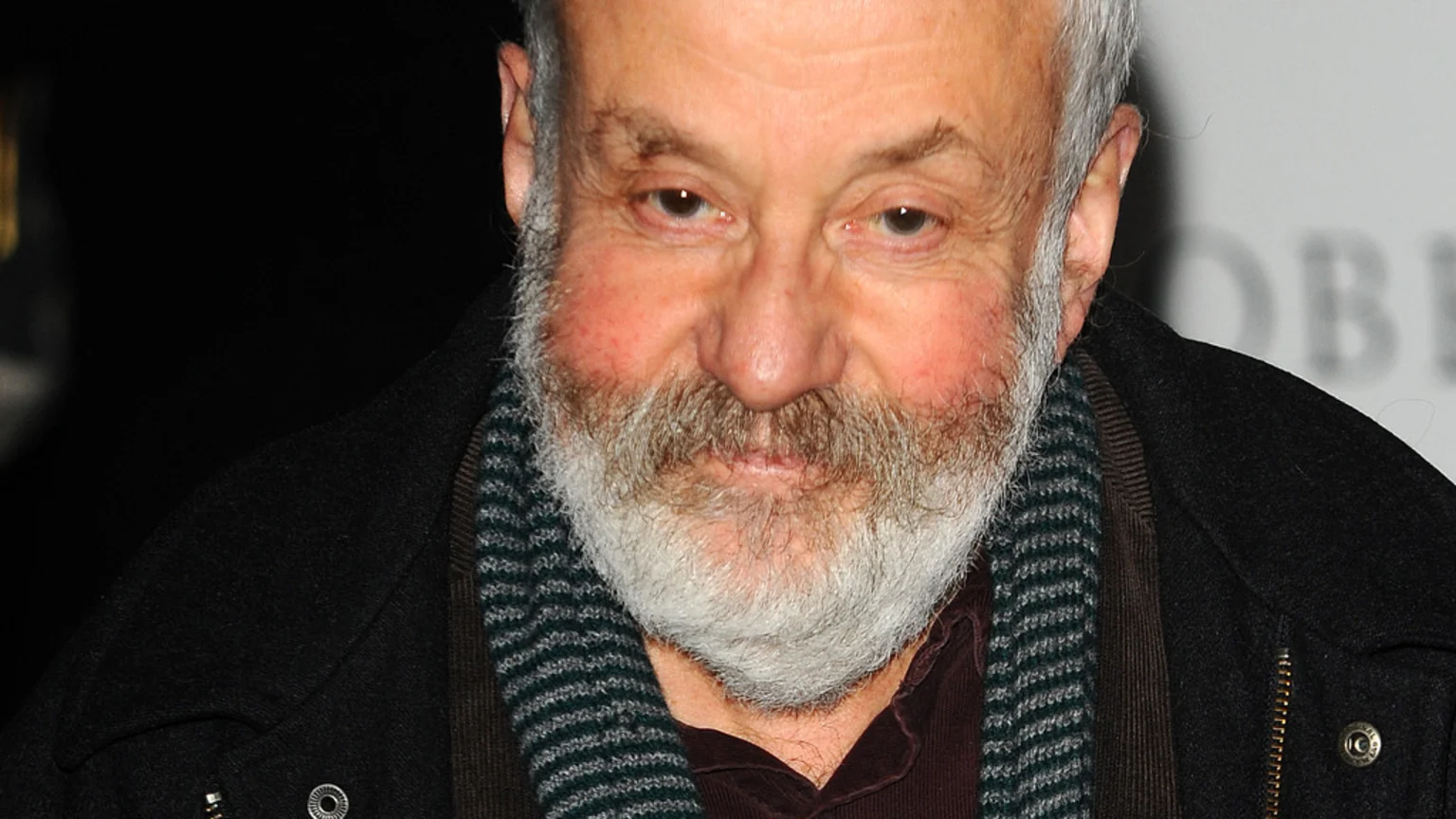 Close-up of filmmaker Mike Leigh, a smiling man with a white beard, grey hair, and rosy cheeks, wearing a dark coat and a striped scarf.