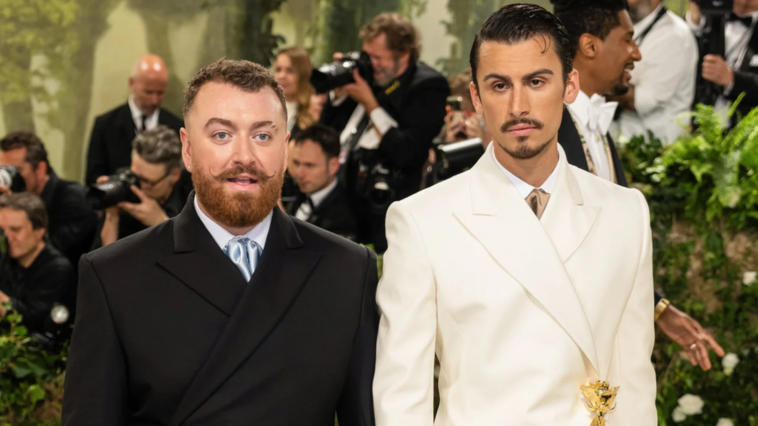 Sam Smith, with a beard and mustache, wearing a dark suit and light blue tie, stands next to Christian Cowan, wearing a white suit with a gold adornment, at an event.