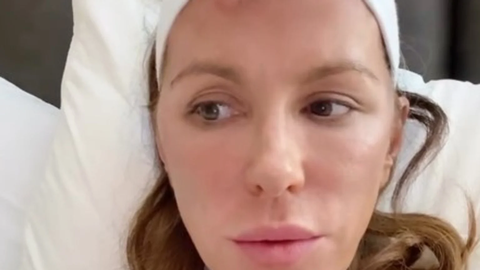 A close-up of Kate Beckinsale lying on a white pillow, wearing a white headband, with brown hair, a small red bump on her forehead, and a pensive expression looking to her left.