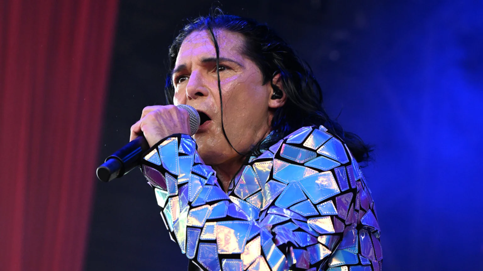 A sweating man with long dark hair and an iridescent, geometric jacket sings into a microphone under blue stage lights.