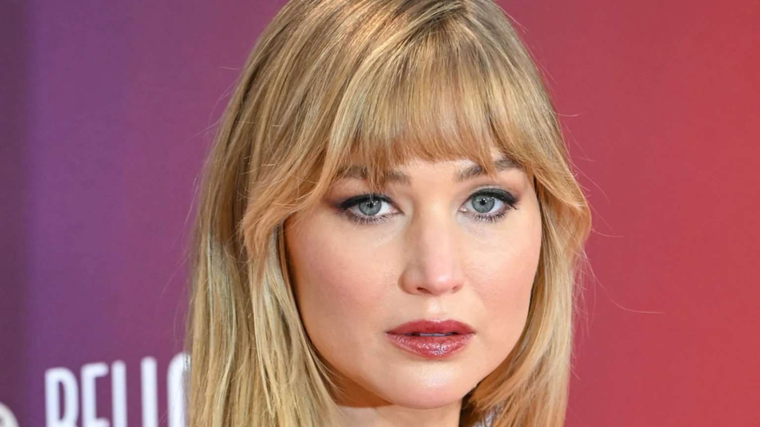 A close-up portrait of Jennifer Lawrence with blonde hair and bangs, blue eyes, and pinkish-red lipstick, against a blurred purple and red background.