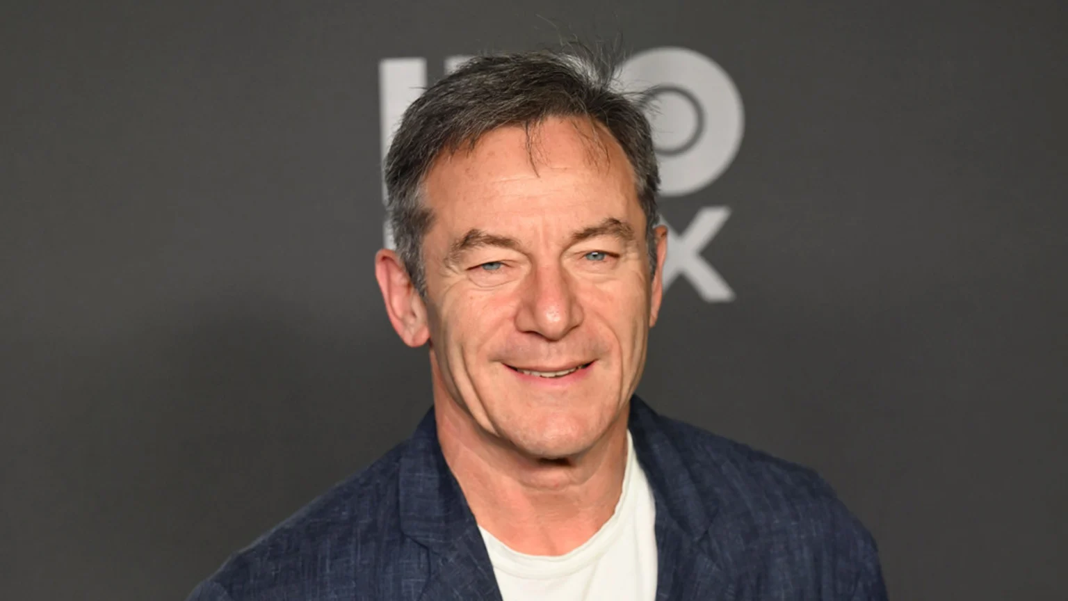 A man with short grey hair and blue eyes smiles at the camera, wearing a blue patterned jacket over a white t-shirt, against a dark grey background with a white HBO Max logo.