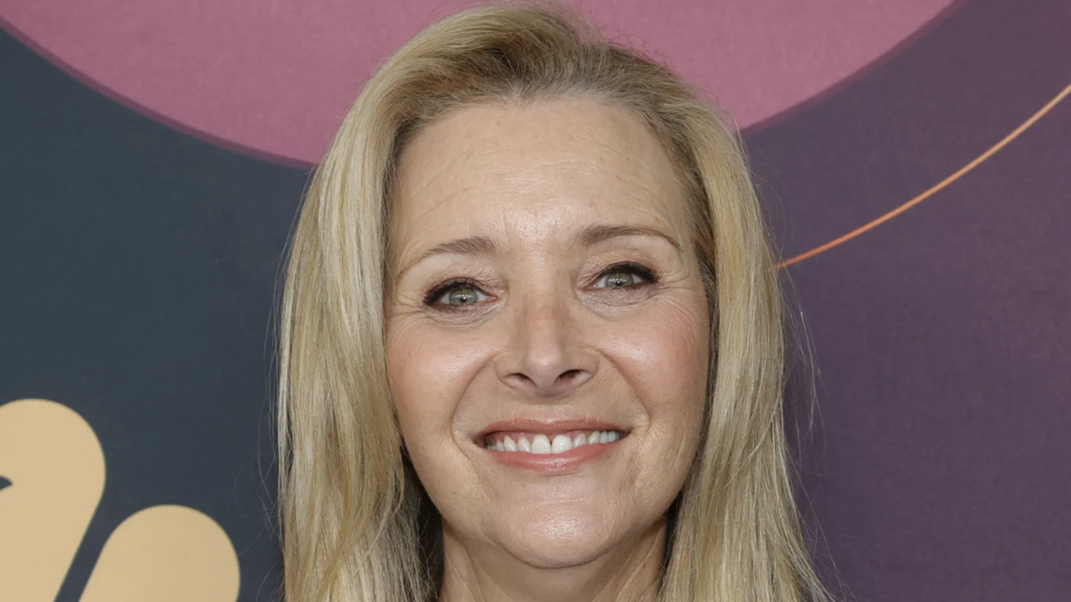 Close-up of Lisa Kudrow smiling, with blonde hair and light makeup, against a blurred, abstract background in shades of dark grey and pinkish-purple.