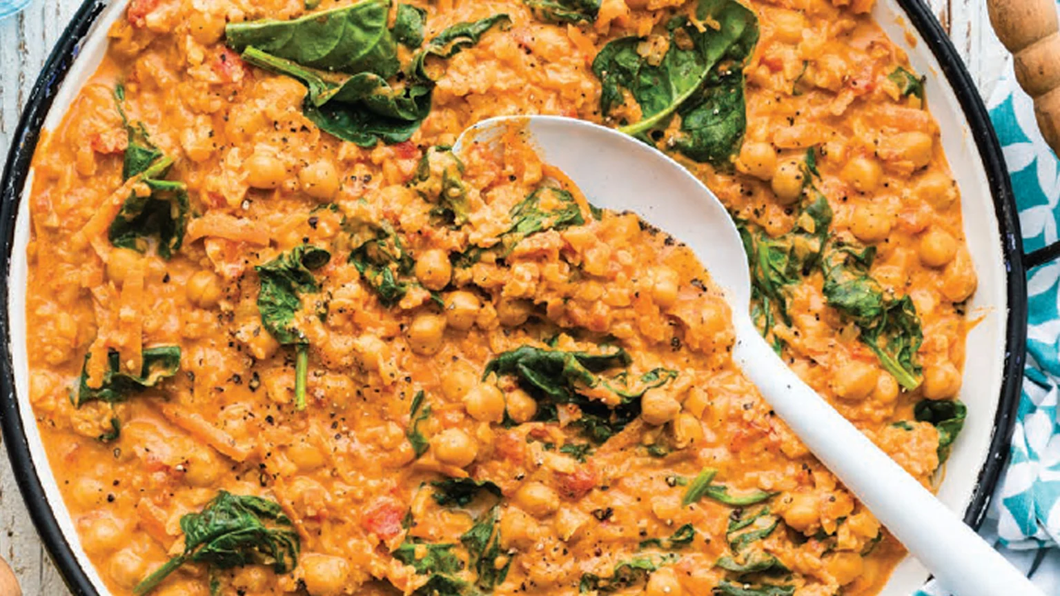 A close-up, overhead view of a creamy, orange-colored chickpea and spinach curry seasoned with black pepper, served in a white dish with a black rim and a white spoon.
