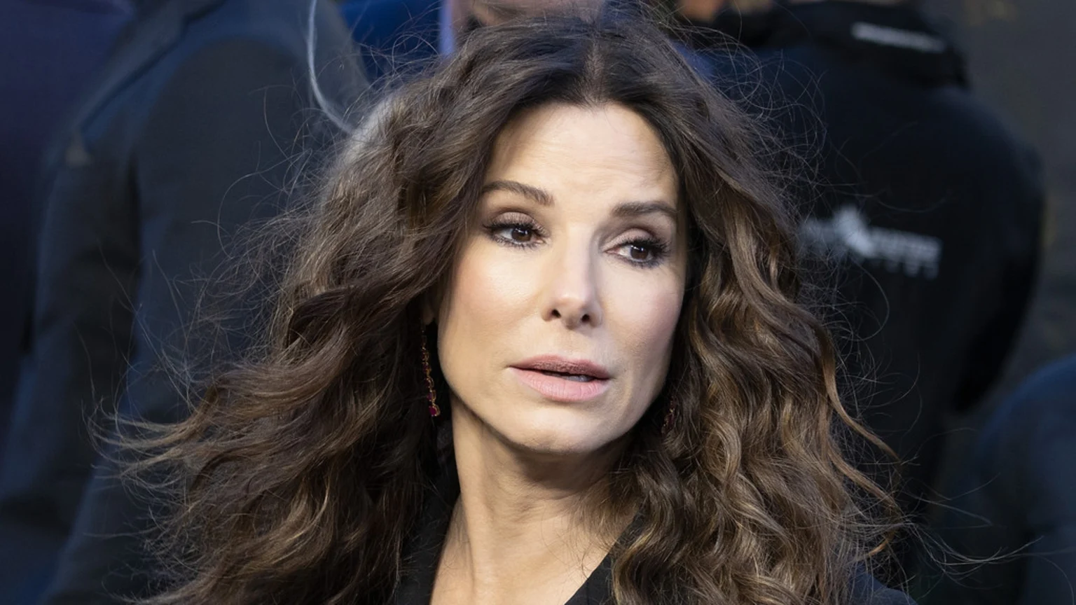 A close-up portrait of actress Sandra Bullock, with long, wavy dark brown hair, subtle makeup, and drop earrings, looking pensively to the side.