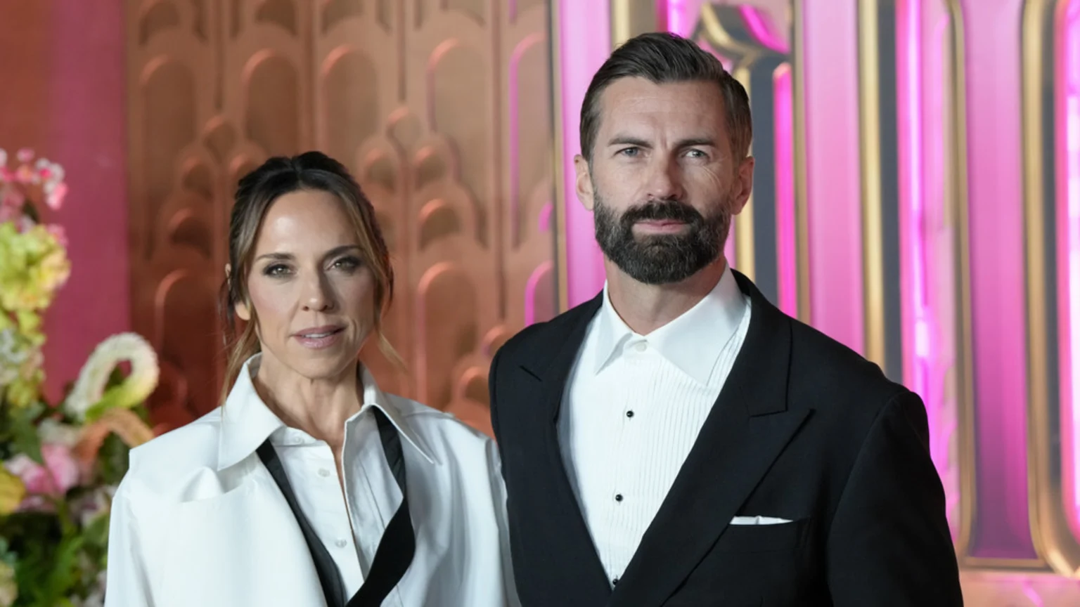 A woman in a white collared shirt and a bearded man in a black suit pose, looking at the camera, against a pink and gold Art Deco-style background with blurred flowers.