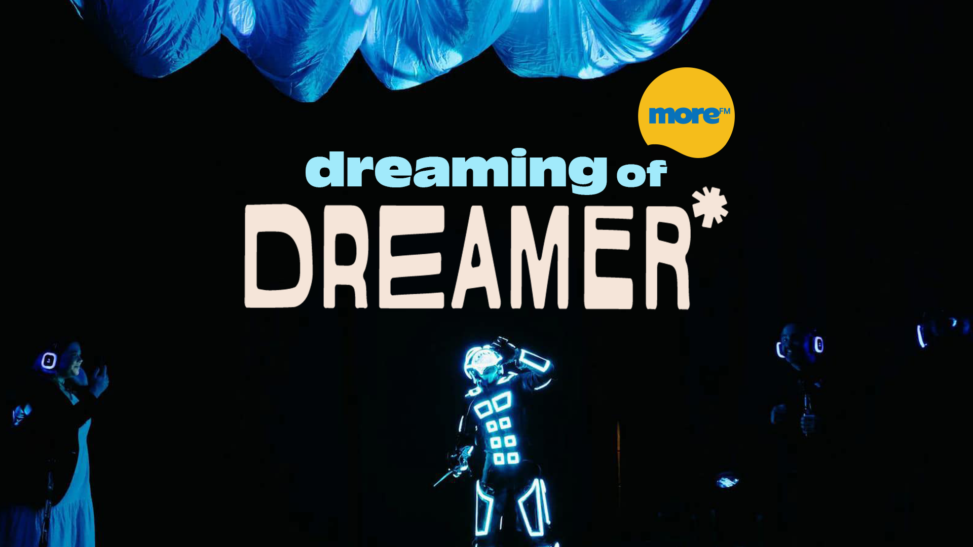 Three performers on a dark stage, one in a glowing futuristic suit, two with glowing headphones, beneath text "dreaming of DREAMER*" and a "more FM" logo, with blue flowing fabric overhead.
