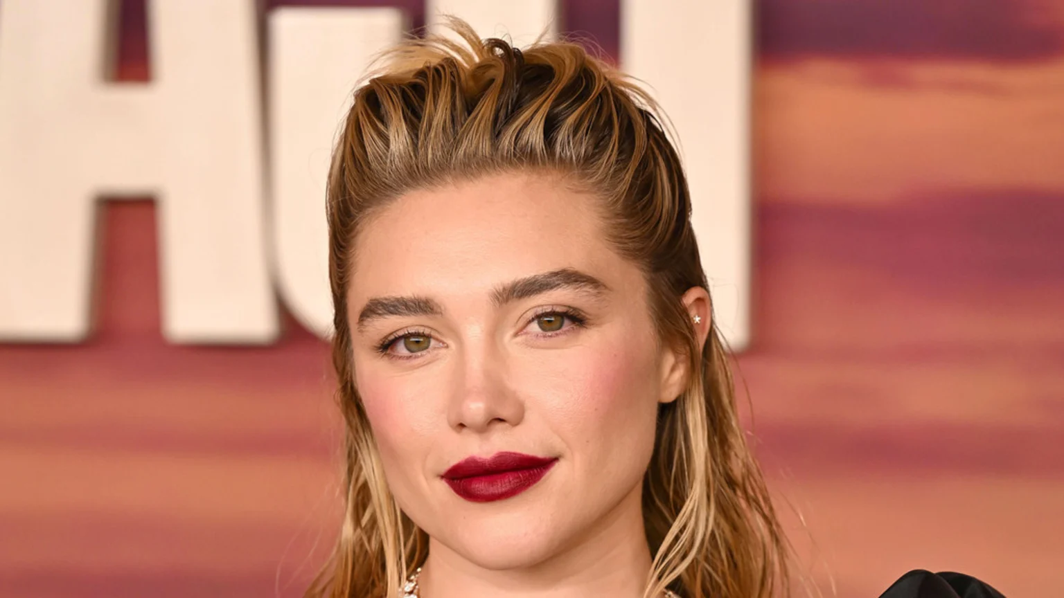 Close-up portrait of actress Florence Pugh with light brown hair pulled back, dark red lipstick, and a star earring in her left ear.