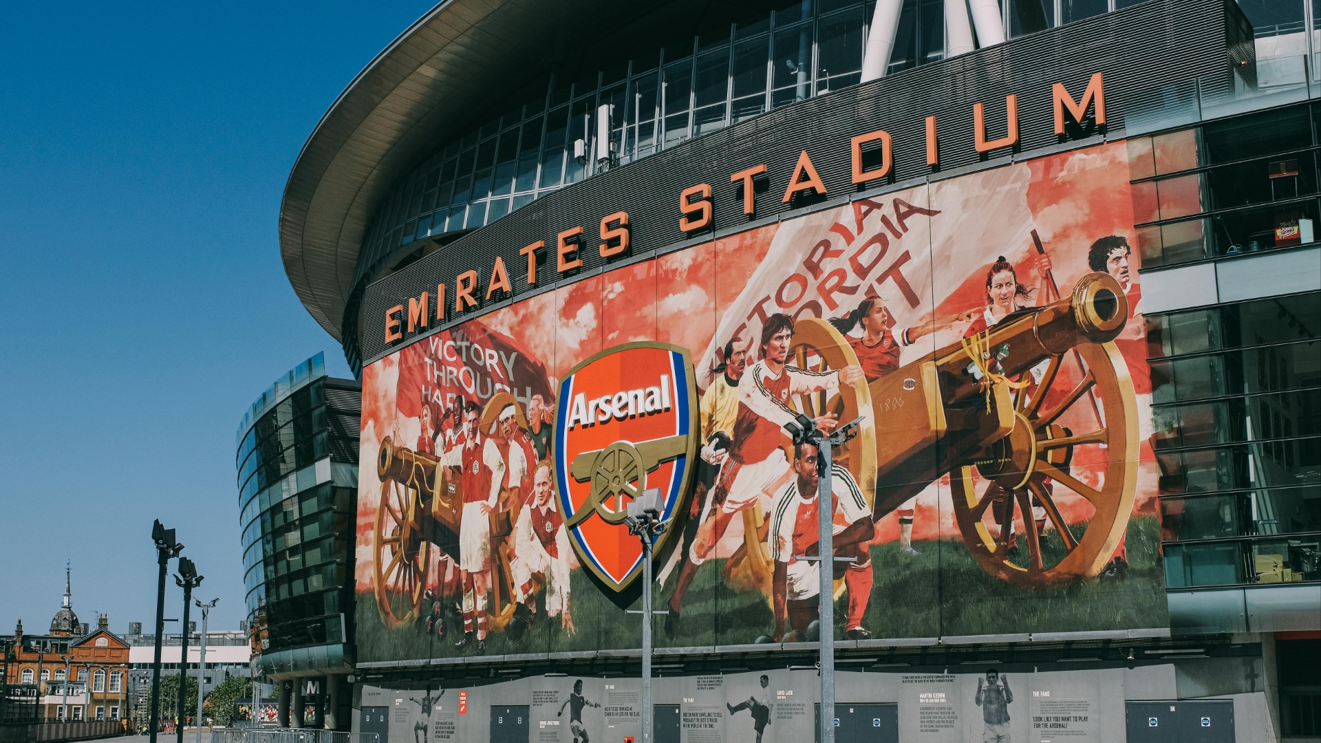 The exterior of Emirates Stadium under a blue sky, featuring a large banner with the Arsenal logo, a cannon, and historical players in red and white kits.