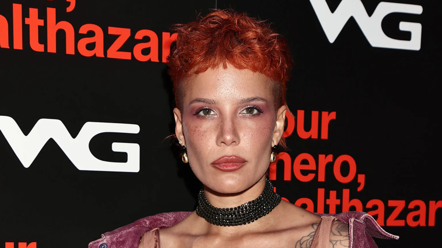 A person with short, reddish-orange hair, freckles, pink eyeshadow, and a black choker looks directly at the camera against a black background with red text and a white "WG" logo.