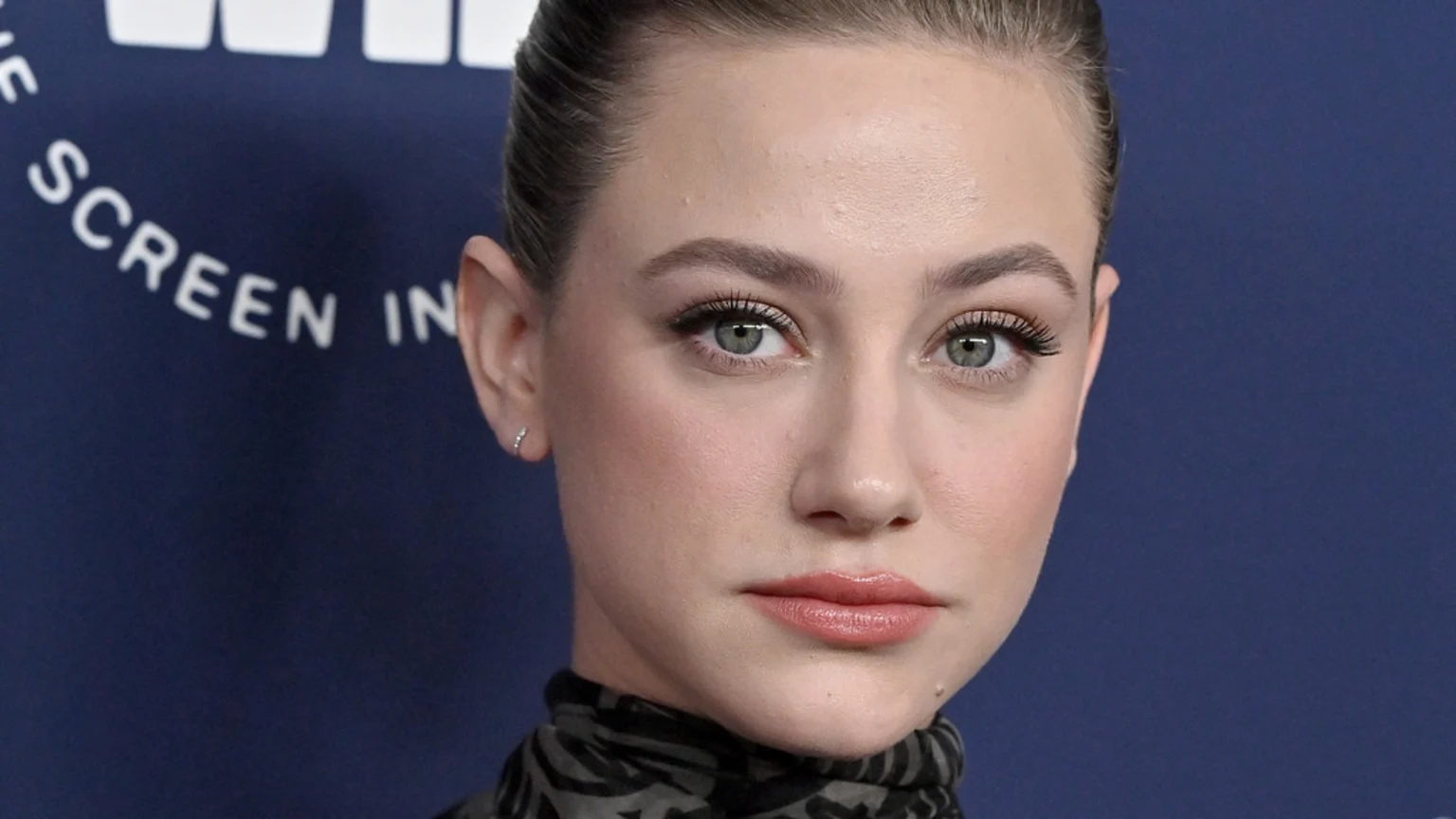 Close-up portrait of Lili Reinhart looking directly at the camera, wearing light makeup, stud earrings, and a patterned high-neck top, against a dark blue background with white text.