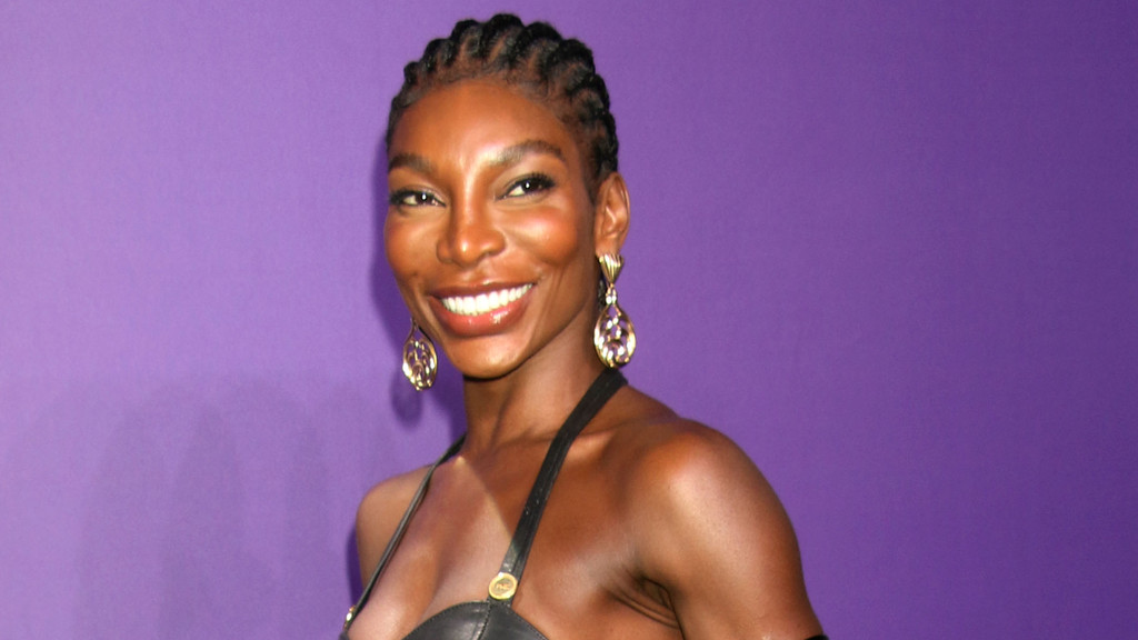 Michaela Coel, a smiling dark-skinned woman with braided hair, wears a black top and large gold drop earrings, pictured against a solid purple background.