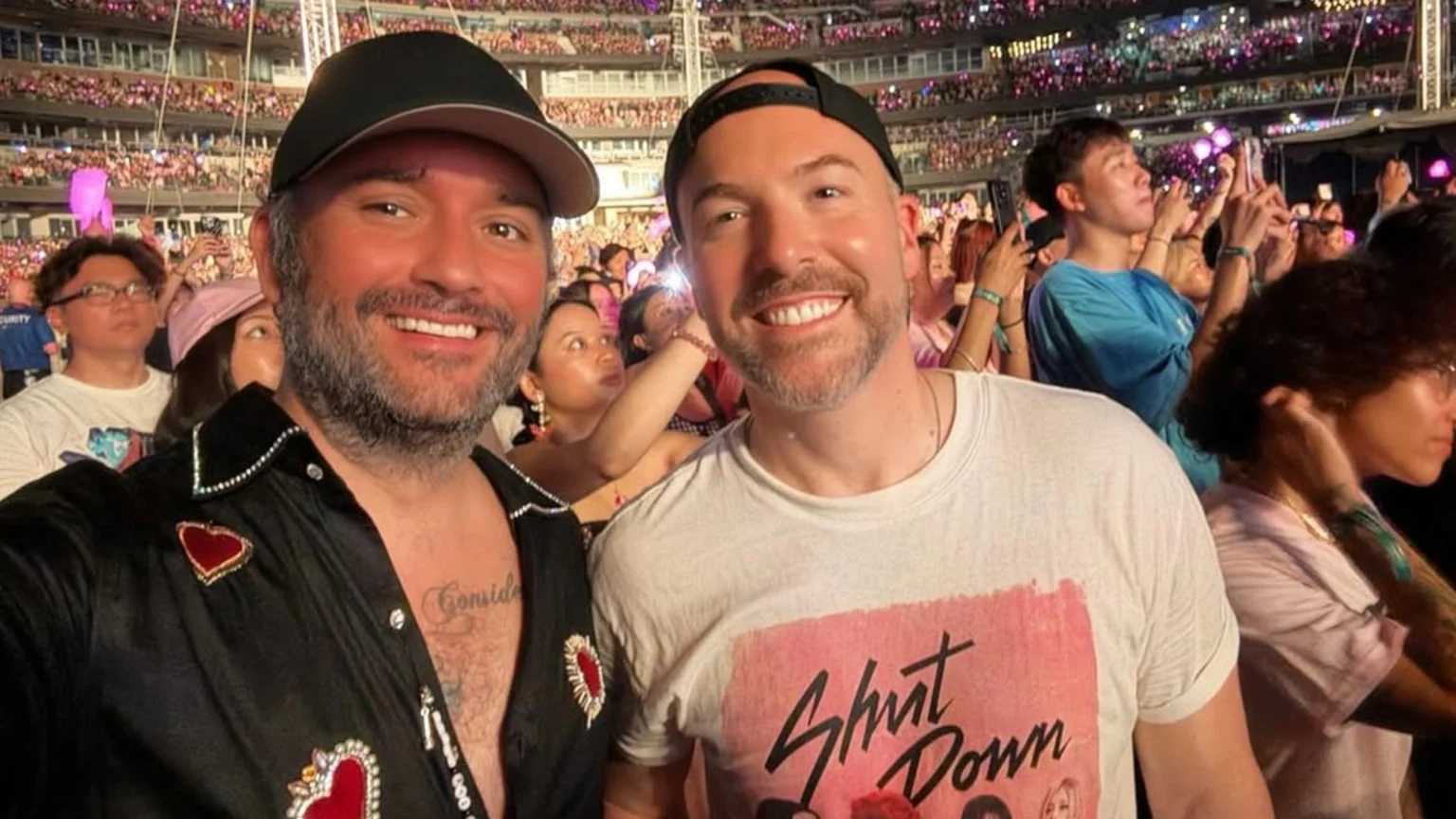 Two smiling men pose for a selfie at a crowded night concert, with one wearing a black shirt with heart designs and the other a white "Shut Down" t-shirt, surrounded by fans holding up phones and light sticks.