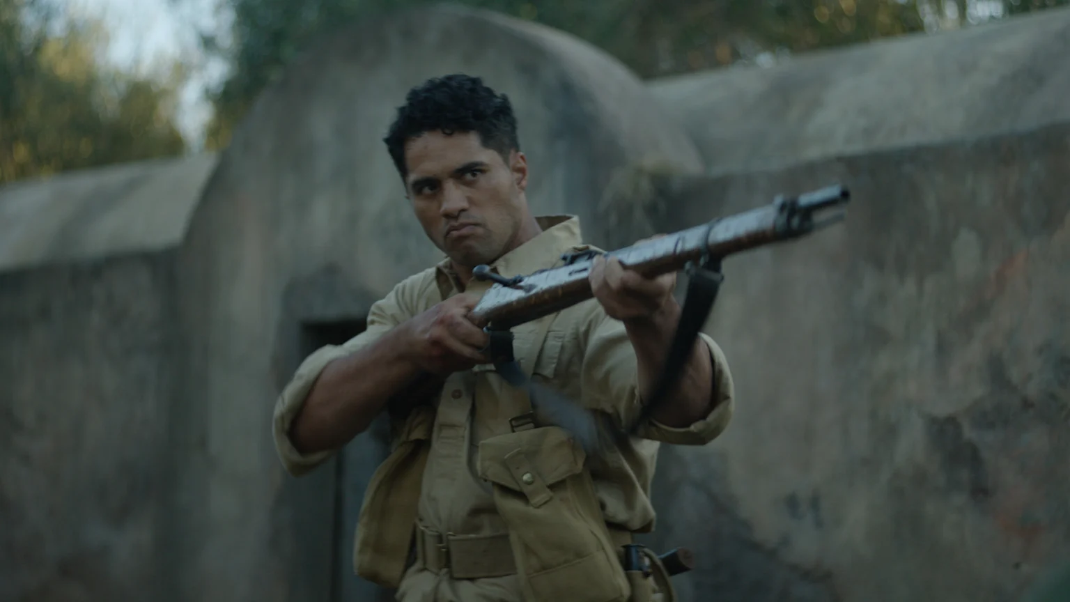 A man with dark, curly hair, dressed in a khaki military uniform, holds a rifle with a stern expression, aiming it.