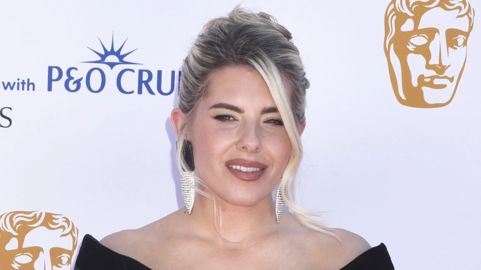 A blonde woman with an updo and large earrings smiles at the camera, wearing an off-the-shoulder black top, with P&O Cruises and BAFTA logos visible in the background.