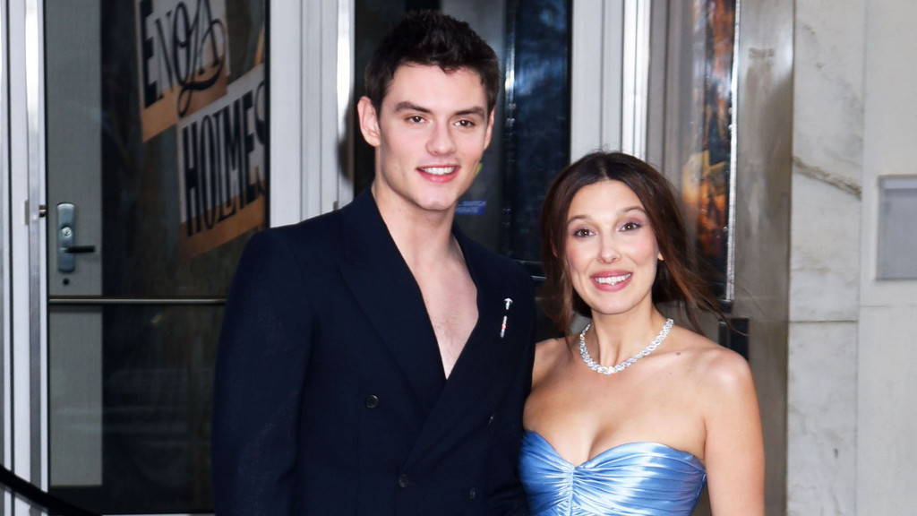 Louis Partridge and Millie Bobby Brown smile at the camera, with Partridge in a dark suit jacket and Brown in a light blue strapless dress and a sparkling necklace, standing in front of glass doors reflecting "ENOLA HOLMES".