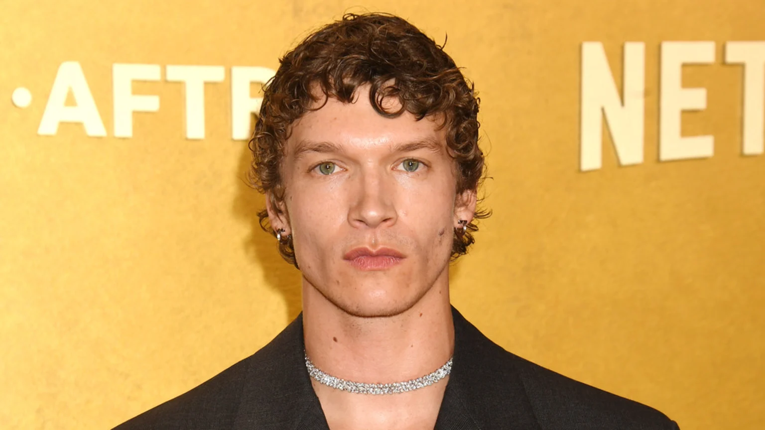 A man identified as Connor Storrie, with curly brown hair, light eyes, and a serious expression, wears a dark jacket and a sparkling silver choker against a yellow background.
