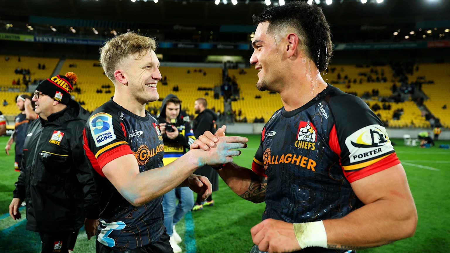 Two smiling Chiefs rugby players in dark jerseys shake hands on a field at night, one player with blue paint on his hand.