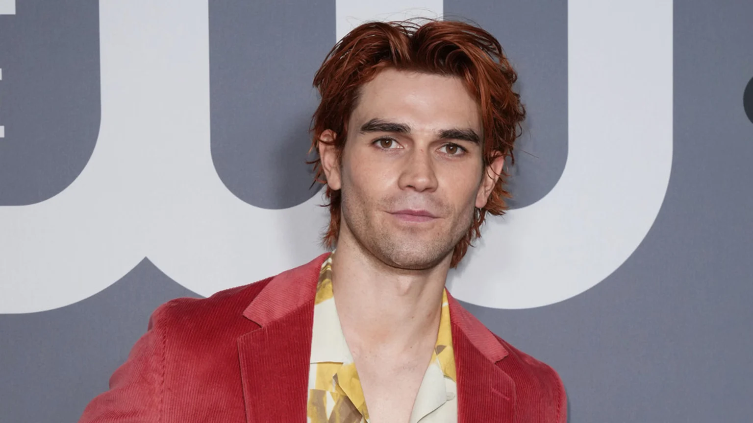A man with reddish-brown hair in a red corduroy blazer and patterned shirt looks directly at the camera against a grey background with the CW logo.