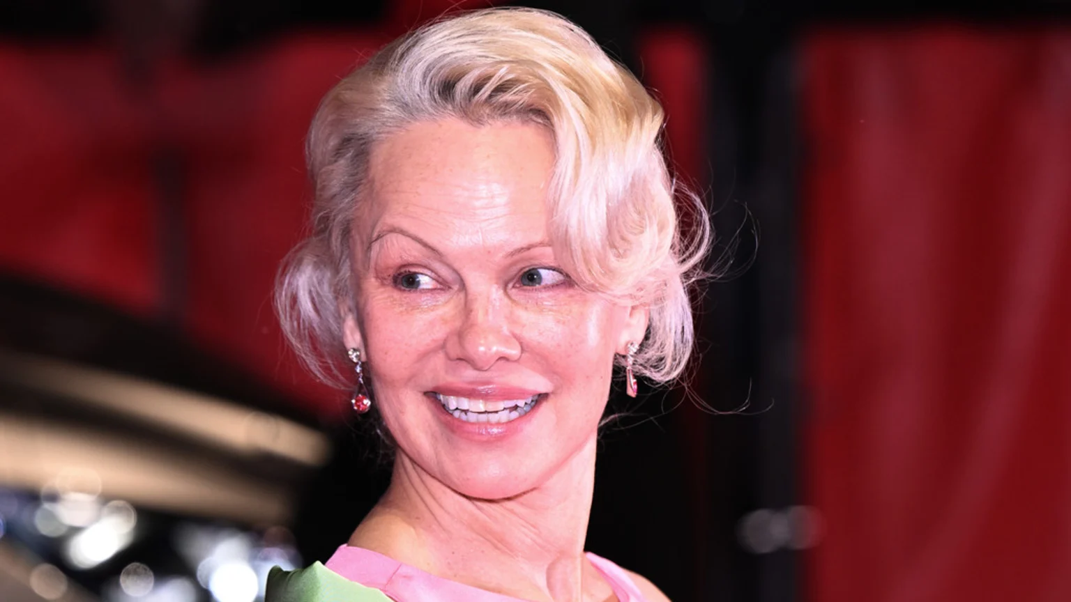 A close-up of Pamela Anderson smiling and looking left, wearing pink earrings and a pink top, with natural blonde hair against a blurry red background.