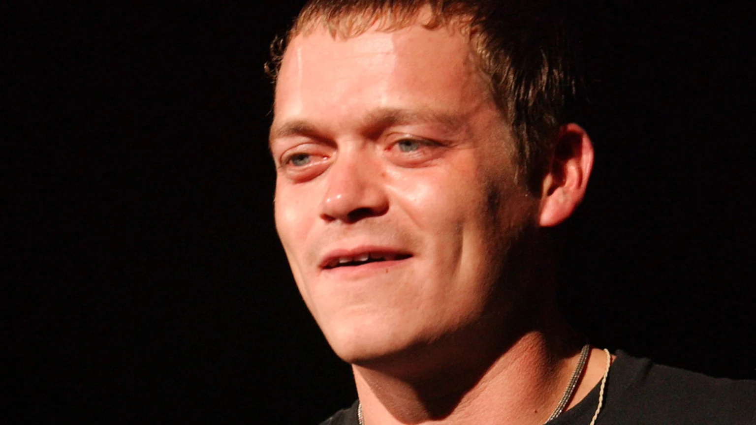 A close-up, head and upper torso portrait of an adult male with light brown hair and blue eyes, wearing a black t-shirt and a silver-colored chain necklace. He looks slightly to the right with a faint smile against a dark, studio-lit background.