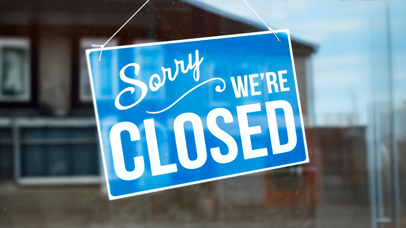 A blue sign displaying "Sorry WE'RE CLOSED" in white text hangs in a window, with a blurred background showing a building and sky.