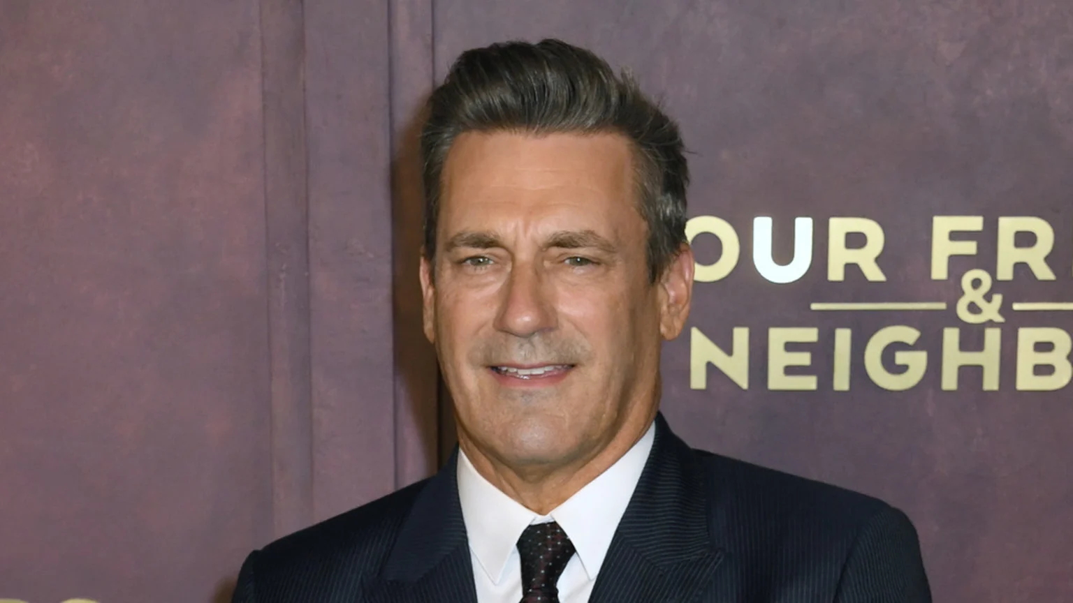 Close-up of actor Jon Hamm smiling slightly, wearing a dark pinstripe suit and patterned tie, against a dark brown background with gold text visible on the right.