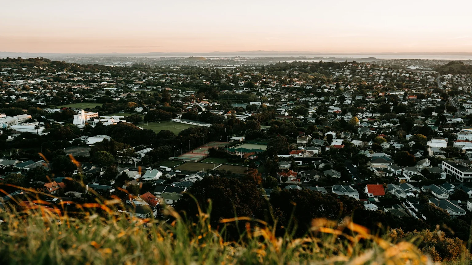 An elevated panoramic view of a dense residential area with numerous houses, green trees, and several sports fields, extending towards a distant body of water under a pale orange sky.