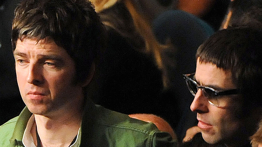 A close-up shows Noel Gallagher on the left in a green shirt, looking down, and Liam Gallagher on the right wearing glasses and looking forward, both with dark hair.