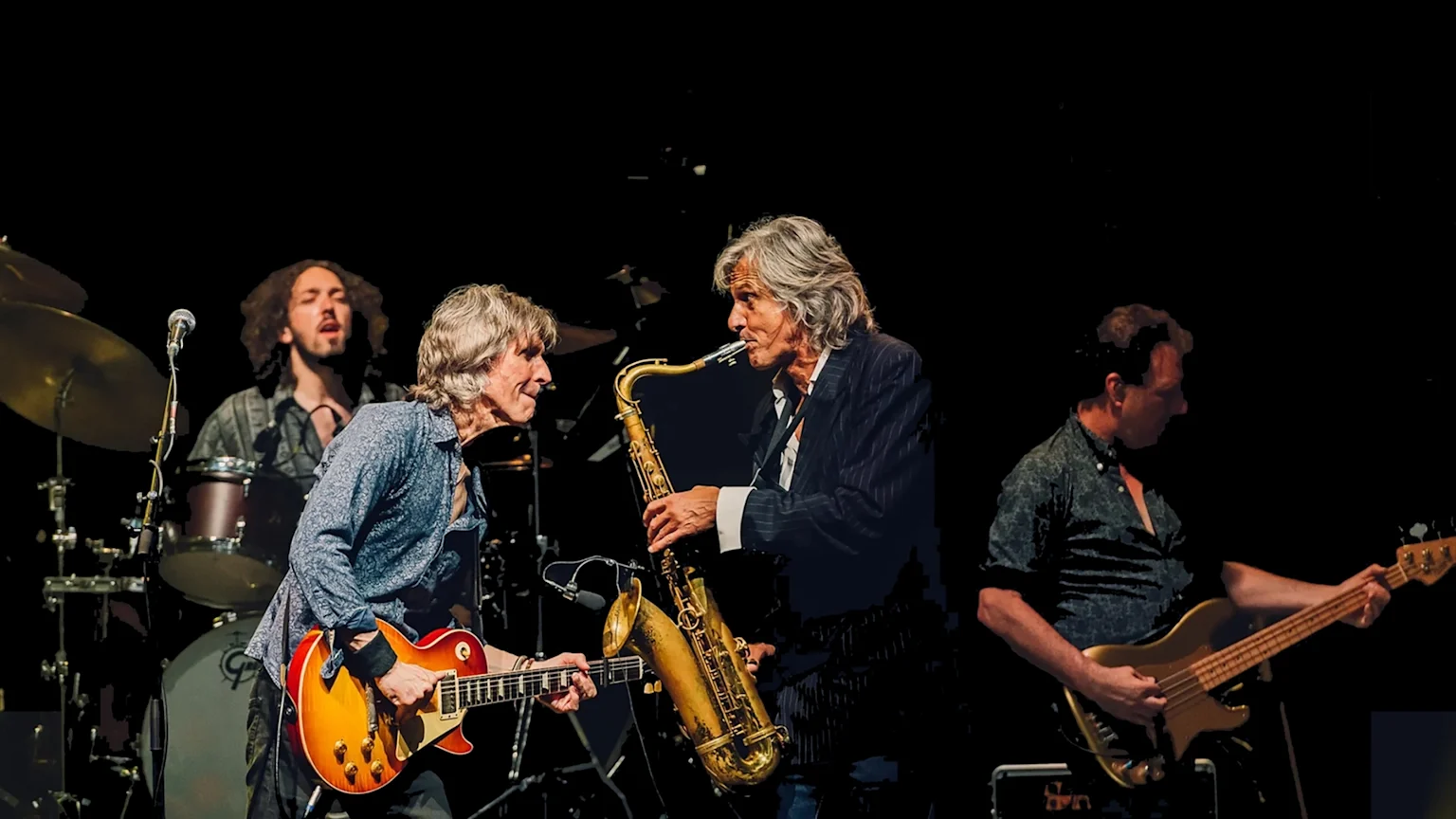 Four musicians perform on a dark stage: a drummer, a guitarist, a saxophonist, and a bassist, with the guitarist and saxophonist in the foreground looking at each other.