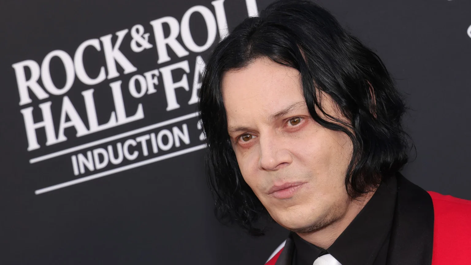 Jack White, with pale skin, black wavy hair, and light brown eyes, wearing a red jacket, poses in front of a "Rock & Roll Hall of Fame Induction" sign.