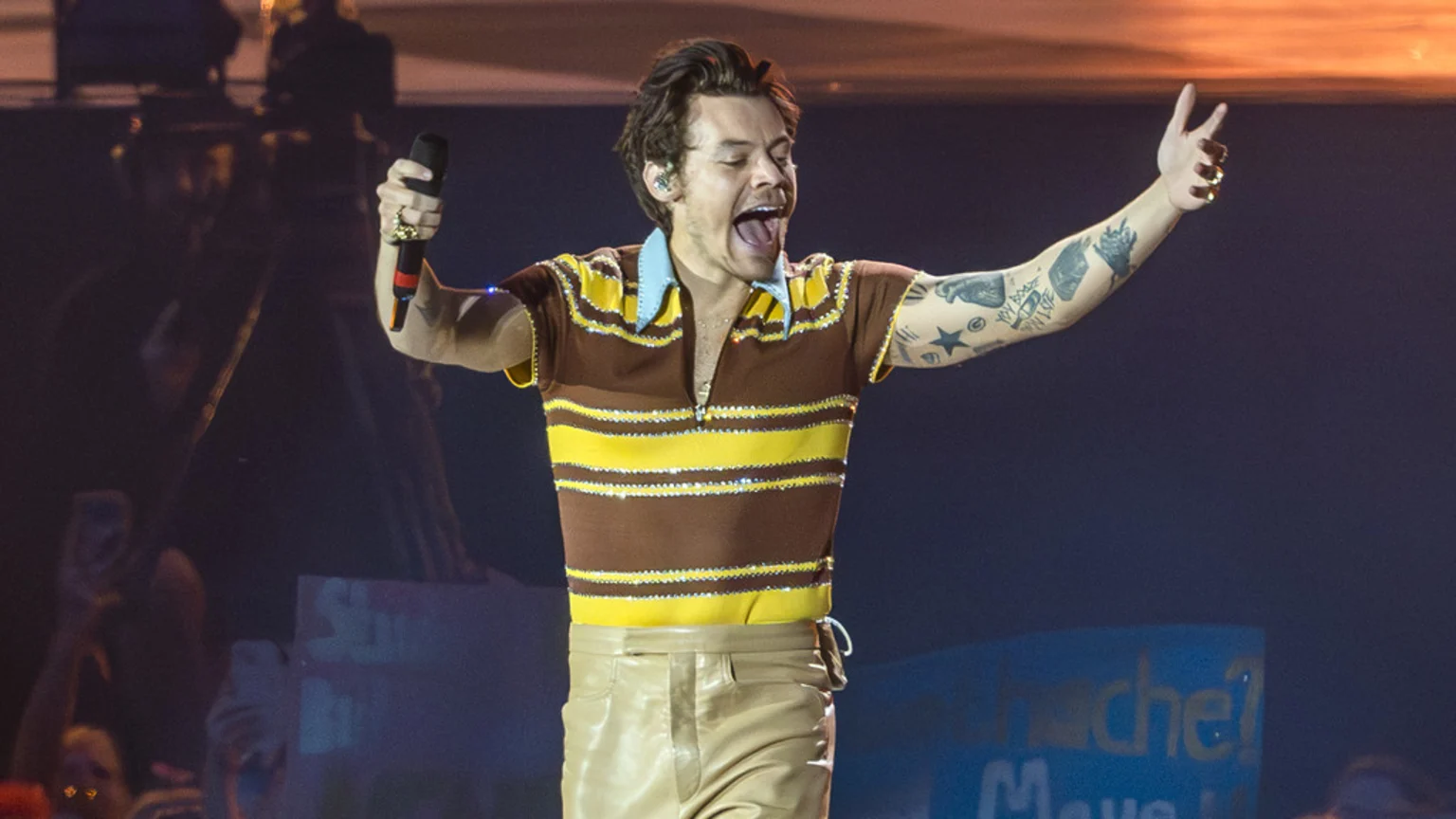 Harry Styles performs on stage, wearing a brown and yellow striped polo shirt, holding a microphone, with his arms wide and mouth open.