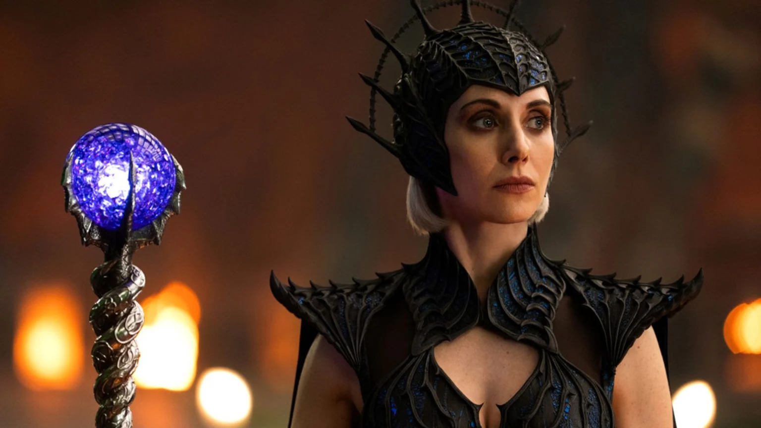 Alison Brie as Evil-Lyn wears a dark, ornate headpiece and matching armor, holding a staff with a glowing blue orb, looking right with a serious expression.