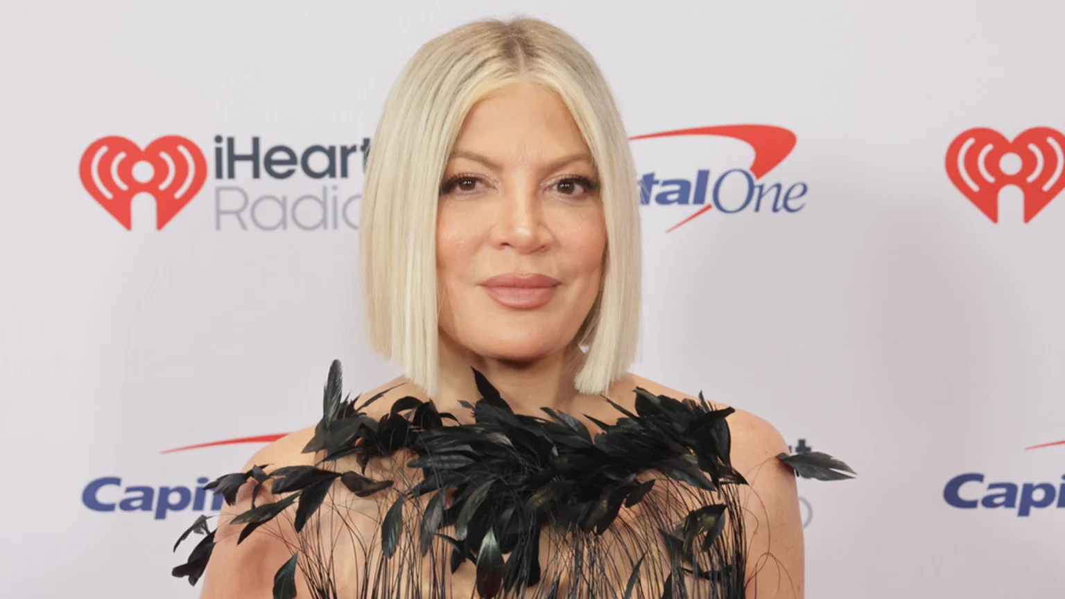 Tori Spelling, a woman with blonde bob hair and light makeup, poses in a black feathered strapless garment against a white backdrop featuring iHeartRadio and CapitalOne logos.
