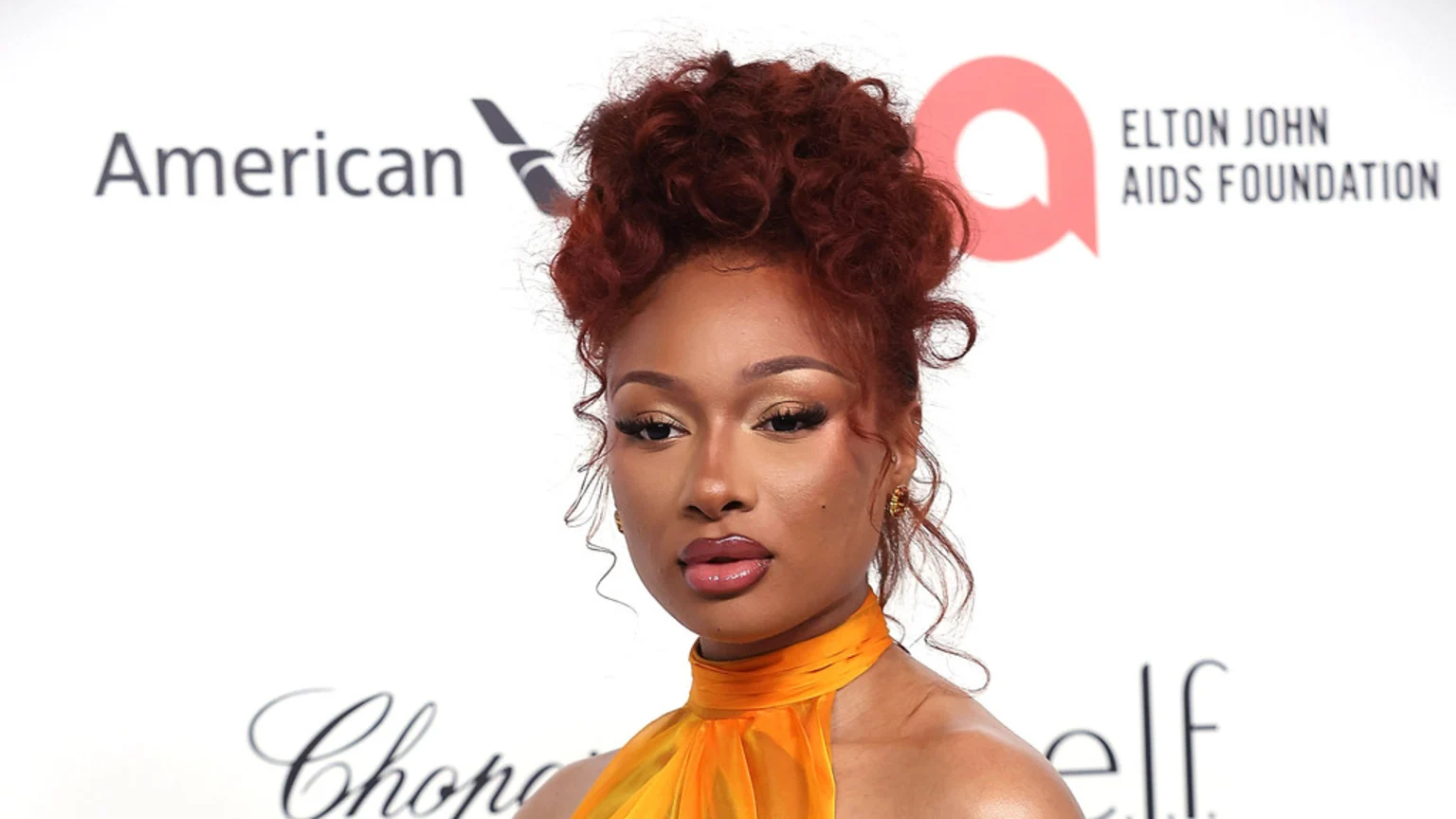 Megan Thee Stallion poses against a white background with sponsor logos, wearing a yellow-orange halter top and her auburn hair in a messy bun.