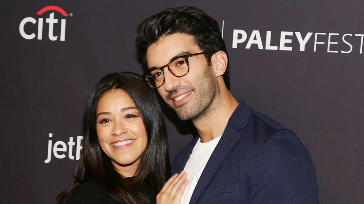 Gina Rodriguez and Justin Baldoni smile and pose closely in front of a dark backdrop with event logos.