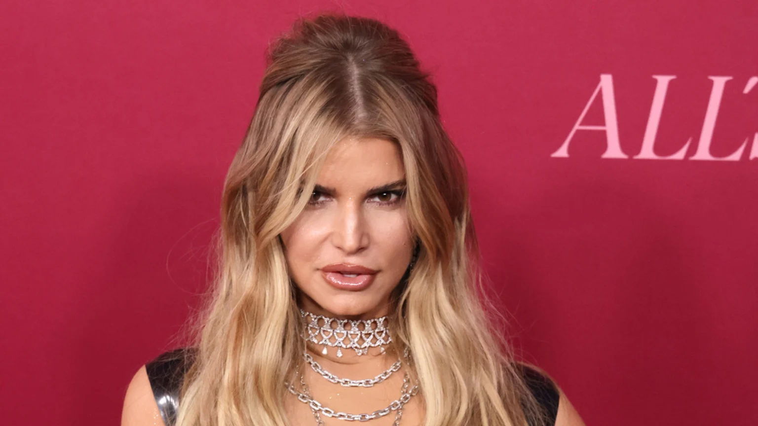 A woman with long blonde hair, styled half-up, looks intensely at the viewer, wearing multiple silver necklaces against a magenta background with partial "ALL" text.