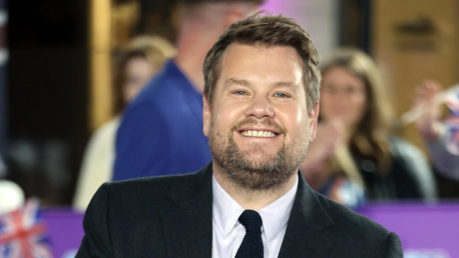 James Corden, a bearded man, smiles broadly at the camera while wearing a dark suit, white shirt, and dark tie, with a blurred audience in the background.